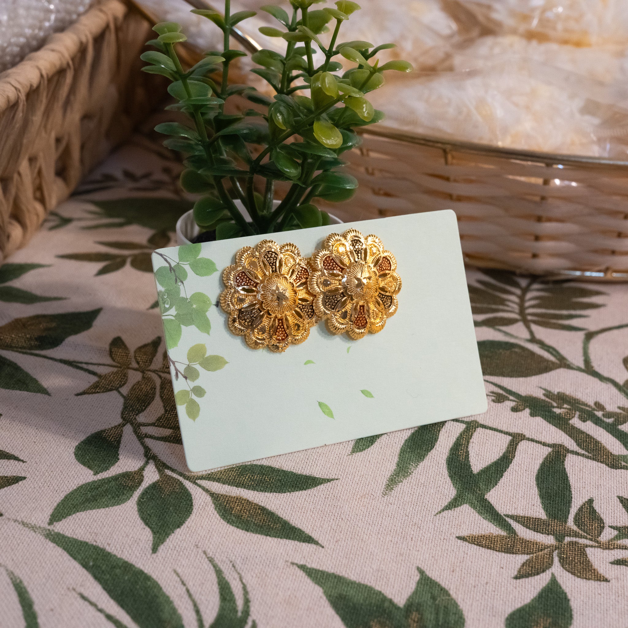 Golden floral earring studs on a card with a plant and woven basket in the background.